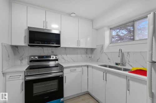 Modern kitchen featuring bright white cabinetry, quartz countertops, a stone-finish backsplash, stainless steel appliances, and a double-basin sink with a pull-down faucet - 1566 Lakewood Road Nw, Edmonton, AB - Indoor Photo Showing Kitchen With Double Sink