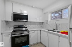 Modern kitchen featuring bright white cabinetry, quartz countertops, a stone-finish backsplash, stainless steel appliances, and a double-basin sink with a pull-down faucet - 