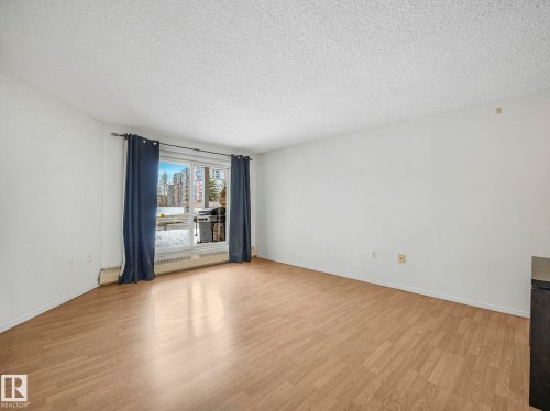 Spacious room featuring wood-finish flooring, white walls, and a large window with blue curtains - 143 10636 120 Street Nw, Edmonton, AB - Indoor Photo Showing Other Room