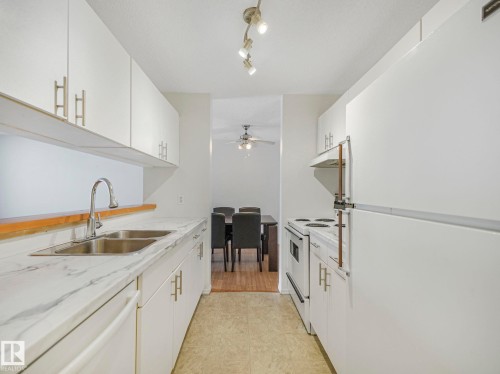 Galley kitchen featuring white cabinetry, a double basin stainless steel sink, and white appliances - 143 10636 120 Street Nw, Edmonton, AB - Indoor Photo Showing Kitchen With Double Sink