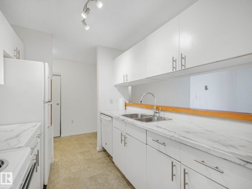 Kitchen featuring white cabinetry, light-toned countertops with veining, a double stainless steel sink with a high-arc faucet, a white dishwasher, and track lighting - 143 10636 120 Street Nw, Edmonton, AB - Indoor Photo Showing Kitchen With Double Sink