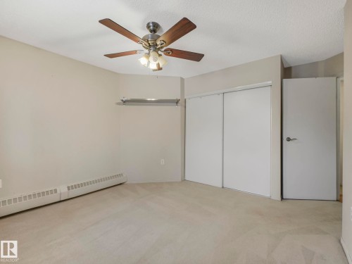 Carpeted interior space featuring a ceiling fan with integrated lighting, neutral wall paint, and white sliding closet doors - 143 10636 120 Street Nw, Edmonton, AB - Indoor Photo Showing Other Room