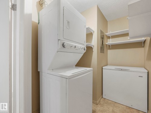 Laundry area featuring a stackable washer and dryer unit, multiple wall-mounted shelves, and a chest freezer - 143 10636 120 Street Nw, Edmonton, AB - Indoor Photo Showing Laundry Room