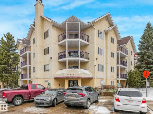 Multi-story building featuring stucco exterior, multiple balconies with purple railings, a prominent central entrance canopy, and a pitched roofline - 143 10636 120 Street Nw, Edmonton, AB - Outdoor With Facade