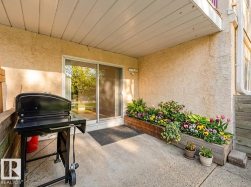 Private concrete patio featuring a sliding glass door, overhead covering, and a built-in planter box with various flowering plants - 143 10636 120 Street Nw, Edmonton, AB - Outdoor With Deck Patio Veranda With Exterior