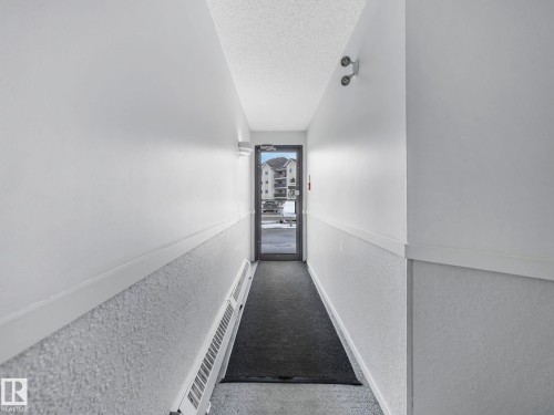 Entryway featuring a security camera, white painted walls, textured lower wall panels, and a dark floor mat - 143 10636 120 Street Nw, Edmonton, AB - Indoor Photo Showing Other Room