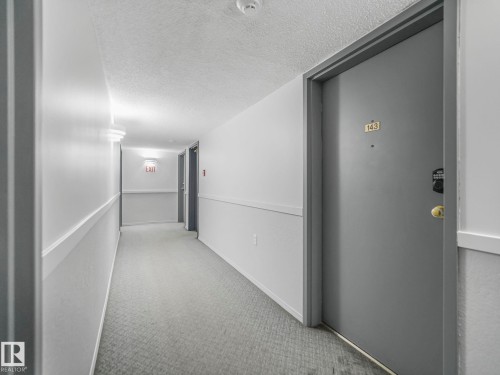 Carpeted hallway featuring a neutral color palette, wall sconce lighting, and a textured ceiling - 143 10636 120 Street Nw, Edmonton, AB - Indoor