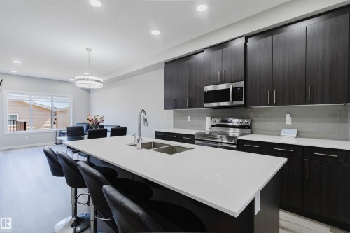 Modern kitchen featuring dark wood-finish cabinetry, stainless steel appliances, a white countertop island with a double sink, and light wood-finish flooring - 343 Edgemont Drive, Edmonton, AB - Indoor Photo Showing Kitchen With Double Sink With Upgraded Kitchen