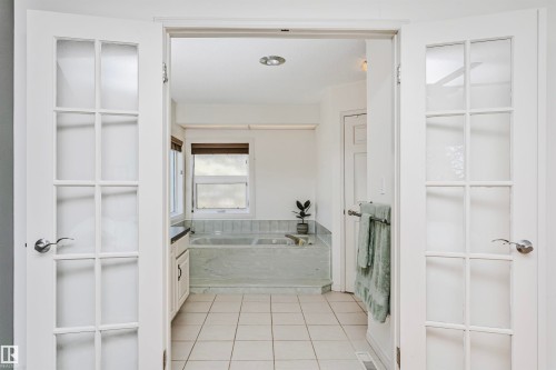 French doors with divided lites opening into a bathroom featuring a built-in tub with stone surround, a window with a privacy blind, and square tile flooring - 17820 57 Avenue, Edmonton, AB - Indoor