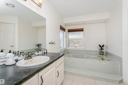 Bathroom featuring a built-in soaking tub with surround tiling, dual windows with blinds, a vanity with a dark countertop, an integrated sink, and an overhead mirror with task lighting - 17820 57 Avenue, Edmonton, AB - Indoor Photo Showing Bathroom