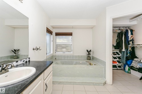 White bathroom featuring a built-in tub with surrounding tile, dual windows with wood-finish blinds, a vanity with a white basin sink, dark countertop, and decorative backsplash, and ceramic tile flooring - 17820 57 Avenue, Edmonton, AB - Indoor Photo Showing Bathroom