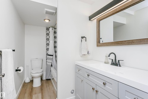 Bathroom featuring a light gray vanity with white countertop, framed mirror with integrated lighting, matte black faucet, and wood-finish flooring - 17820 57 Avenue, Edmonton, AB - Indoor Photo Showing Bathroom