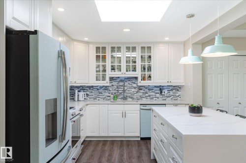 Modern kitchen featuring an expansive center island with an integrated countertop, white cabinetry with glass-front uppers, a mosaic tile backsplash, and wood-finish flooring - 10339 36A Avenue, Edmonton, AB - Indoor Photo Showing Kitchen With Upgraded Kitchen