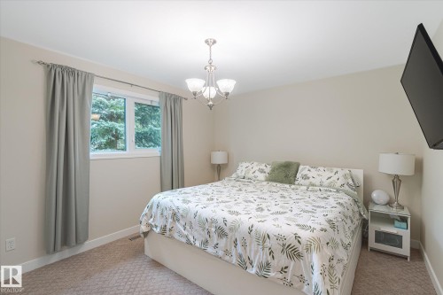 Bright bedroom featuring neutral wall tones, a large window, a ceiling-mounted chandelier, wall-to-wall carpeting, and a wall-mounted television - 10339 36A Avenue, Edmonton, AB - Indoor Photo Showing Bedroom
