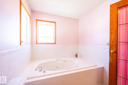White tiled corner soaking tub with chrome fixtures, two windows with wood trim, and a wood-paneled door - 5736 161 Avenue, Edmonton, AB - Indoor Photo Showing Bathroom