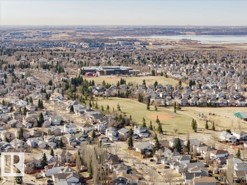 Expansive aerial showcasing a residential neighborhood with numerous detached homes, mature trees, open green spaces including a baseball diamond, and a body of water in the distance - 7 Doucette Place, St. Albert, AB - Outdoor With View