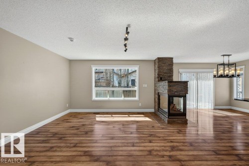 Spacious room featuring wood-finish flooring, a contemporary stone fireplace with a three-sided firebox, a white-framed window, and a modern chandelier - 10 Erickson Close, St. Albert, AB - Indoor With Fireplace