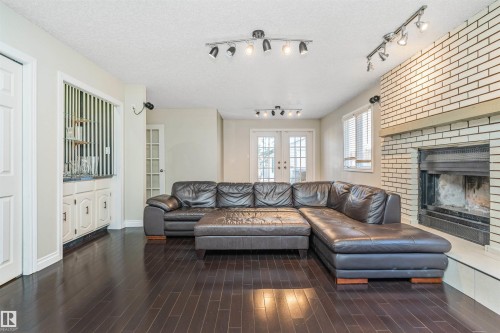 Living area featuring dark wood-finish flooring, a white brick fireplace with a wood mantel, and track lighting fixtures - 6016 177 Street, Edmonton, AB - Indoor Photo Showing Living Room With Fireplace