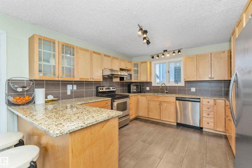 Kitchen featuring warm-toned cabinetry, granite countertops, stainless steel appliances, a tiled backsplash, and wood-finish flooring - 6016 177 Street, Edmonton, AB - Indoor Photo Showing Kitchen With Stainless Steel Kitchen