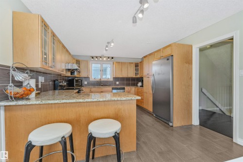 Kitchen featuring wood cabinetry, stone-finish countertops, stainless steel appliances, a ceramic tile backsplash, and wood-finish flooring - 6016 177 Street, Edmonton, AB - Indoor Photo Showing Kitchen
