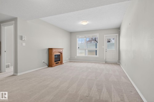 Spacious carpeted room featuring a wood-finish electric fireplace, light gray wall paint, white trim, and a window with frosted lower panes - 34 3003 34 Avenue, Edmonton, AB - Indoor Photo Showing Other Room