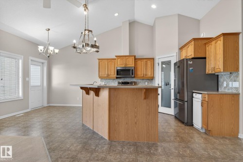 Vaulted ceiling kitchen featuring wood-finish cabinetry, a central island with integrated sink, stainless steel refrigerator, and tiled flooring - 34 3003 34 Avenue, Edmonton, AB - Indoor Photo Showing Kitchen