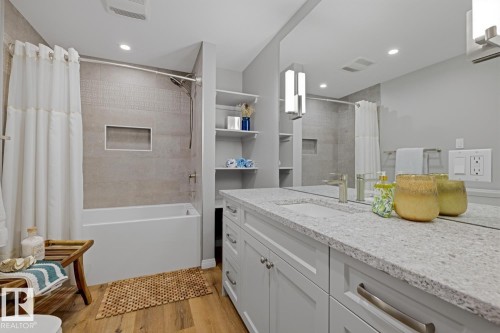 Bathroom featuring a bathtub with tiled surround and built-in niche, wood-finish flooring, full-width vanity mirror, light grey cabinetry, and a light-toned countertop - 17604 85 Avenue, Edmonton, AB - Indoor Photo Showing Bathroom