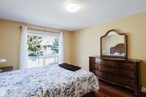 Room featuring warm-toned walls, hardwood flooring, and a large window with white sheer curtains - 9 102 Bridgeport Crossing, Leduc, AB - Indoor Photo Showing Bedroom