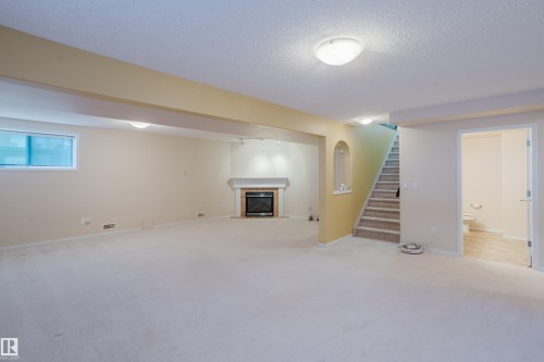Spacious room featuring light-colored carpeting, a fireplace with a mantel, a small rectangular window, and an open doorway leading to a staircase and another room - 9 102 Bridgeport Crossing, Leduc, AB - Indoor Photo Showing Basement