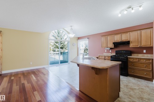 Open concept living space featuring wood-finish flooring, a kitchen island with sink, black range, wood cabinetry, and a sliding glass door with an arched transom leading to an outdoor deck - 9 102 Bridgeport Crossing, Leduc, AB - Indoor Photo Showing Kitchen
