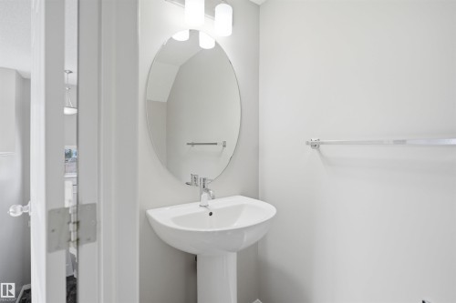 Pedestal sink with chrome faucet, an oval mirror, and wall-mounted light fixture - 33 7385 Edgemont Way Nw, Edmonton, AB - Indoor Photo Showing Bathroom