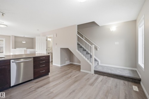 Open-concept living area featuring wood-finish flooring, a kitchen island with stone-finish countertop and stainless steel dishwasher, a carpeted staircase with white risers and black metal spindles, and a recessed wall light fixture - 33 7385 Edgemont Way Nw, Edmonton, AB - Indoor Photo Showing Kitchen