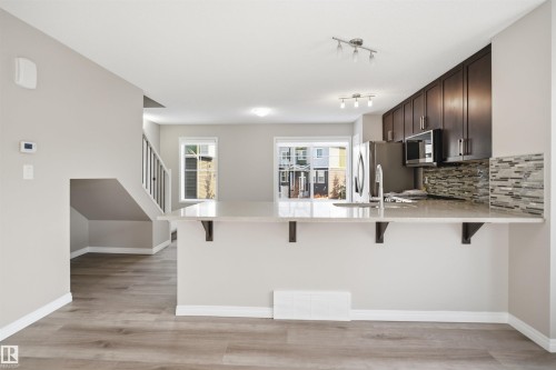 Open-concept kitchen featuring an extended peninsula with light-toned countertop, dark wood-finish cabinetry, stainless steel appliances, and a mosaic tile backsplash - 33 7385 Edgemont Way Nw, Edmonton, AB - Indoor Photo Showing Kitchen With Upgraded Kitchen