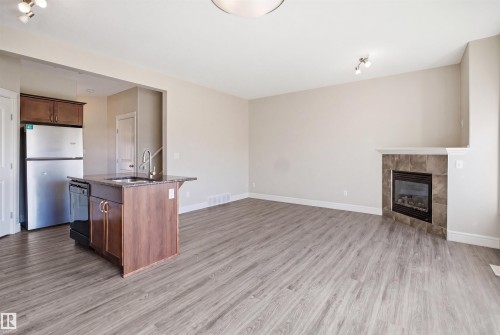 Open-concept living area featuring wood-finish flooring, a kitchen island with built-in sink and stainless steel appliances, and a tiled fireplace with a mantel - 19 15151 43 Street, Edmonton, AB - Indoor With Fireplace