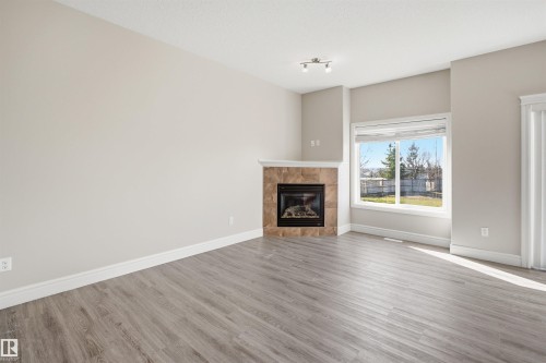 Living area featuring a corner fireplace with a tiled surround, a large window, light-toned walls, wood-finish flooring, and track lighting - 19 15151 43 Street, Edmonton, AB - Indoor Photo Showing Living Room With Fireplace