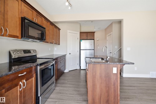 Contemporary kitchen featuring wood-finish cabinetry, dark countertops, and stainless steel appliances including a built-in microwave, range, and refrigerator - 19 15151 43 Street, Edmonton, AB - Indoor Photo Showing Kitchen With Double Sink