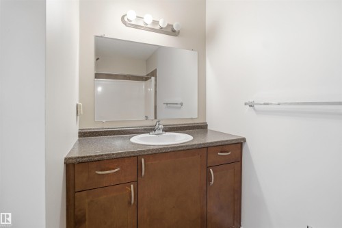 Bathroom featuring a brown wood-finish vanity with a built-in sink, a speckled countertop, an overhead vanity light, and a wall-mounted mirror - 19 15151 43 Street, Edmonton, AB - Indoor Photo Showing Bathroom