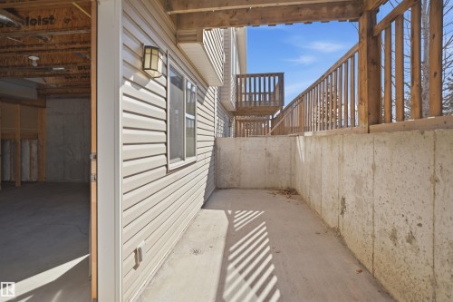 Concrete patio beneath a wood-railed deck, featuring a window and wall-mounted light fixture on the siding - 19 15151 43 Street, Edmonton, AB -  With Exterior