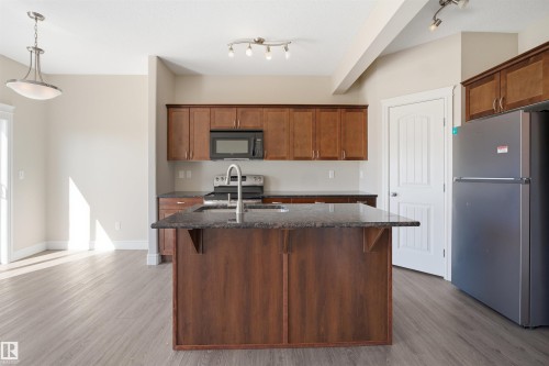 Kitchen featuring a center island with sink, wood-finish cabinetry, dark countertops, and stainless steel appliances - 19 15151 43 Street, Edmonton, AB - Indoor Photo Showing Kitchen With Double Sink