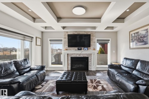 Living area featuring a coffered ceiling with recessed lighting, wood-finish flooring, a stone-clad fireplace with a decorative mantel, and expansive windows - 731 57 Street Sw, Edmonton, AB - Indoor Photo Showing Living Room With Fireplace
