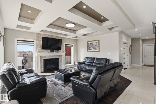Living area featuring a stone-clad fireplace with a white mantel, tray ceilings with recessed lighting, wood-finish flooring, and large windows - 731 57 Street Sw, Edmonton, AB - Indoor Photo Showing Living Room With Fireplace