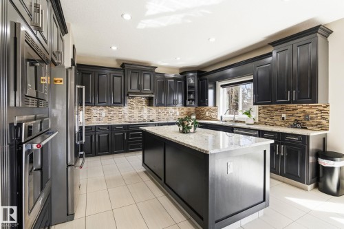 Kitchen featuring dark wood cabinetry, a central island with a light-toned countertop, stainless steel appliances, a mosaic tile backsplash, and light-toned tile flooring - 731 57 Street Sw, Edmonton, AB - Indoor Photo Showing Kitchen With Upgraded Kitchen