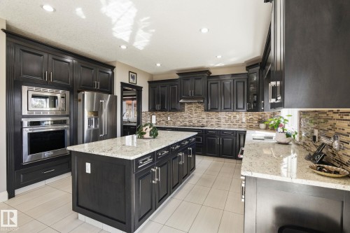 Kitchen featuring dark cabinetry, stainless steel appliances, a center island with a light-toned countertop, and a mosaic tile backsplash - 731 57 Street Sw, Edmonton, AB - Indoor Photo Showing Kitchen With Upgraded Kitchen