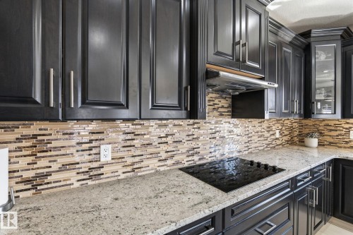 Kitchen featuring dark wood-finish cabinetry with silver-tone hardware, a mosaic tile backsplash, light-colored speckled countertops, and an integrated black cooktop - 731 57 Street Sw, Edmonton, AB - Indoor Photo Showing Kitchen With Upgraded Kitchen
