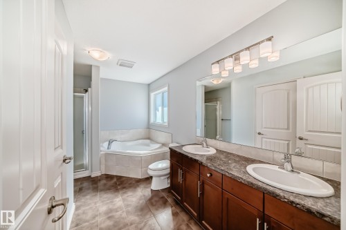 Bathroom featuring a dual-sink vanity with a dark wood-finish cabinet and stone-look countertop, a corner soaking tub with tiled surround, a glass-enclosed shower, and tiled flooring - 6723 19 Avenue, Edmonton, AB 