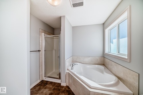 Bathroom featuring a built-in corner soaking tub with tiled surround, a separate glass-enclosed shower, a window providing natural light, a ceiling-mounted light fixture, and dark-toned tile flooring - 6723 19 Avenue, Edmonton, AB 