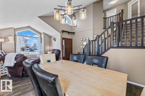 Open-concept dining area with a modern chandelier, vaulted ceilings, wood-finish flooring, and a prominent staircase with dark wood railings - 86 Westpark Way, Fort Saskatchewan, AB - Indoor Photo Showing Dining Room