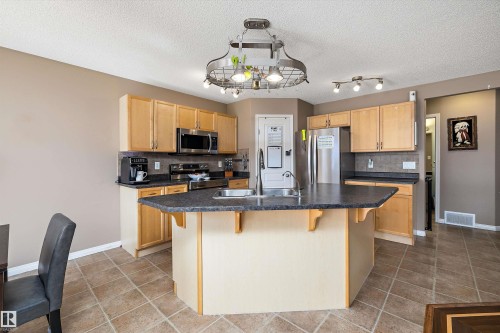 Kitchen featuring a central island with a sink, wood-finish cabinetry, stainless steel appliances, dark countertops, and ceramic tile flooring - 992 Regency Drive, Sherwood Park, AB - Indoor Photo Showing Kitchen With Double Sink