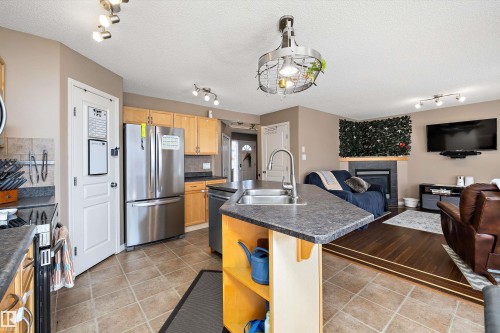 Open-concept kitchen and living area featuring stainless steel appliances, a kitchen island with an integrated sink, wood-finish cabinetry, ceramic tile flooring, and a fireplace with a dark mantel - 992 Regency Drive, Sherwood Park, AB - Indoor Photo Showing Kitchen With Double Sink