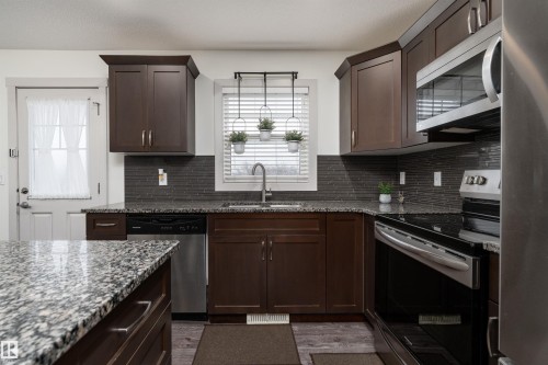 Kitchen featuring dark wood cabinetry, granite countertops, a dark tile backsplash, stainless steel appliances, and wood-finish flooring - 15 5203 149 Avenue, Edmonton, AB - Indoor Photo Showing Kitchen With Upgraded Kitchen